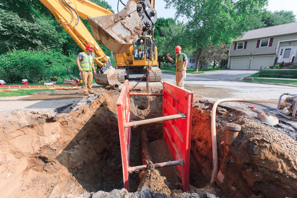 Two men helping lower shoring with an excavator into a hole.